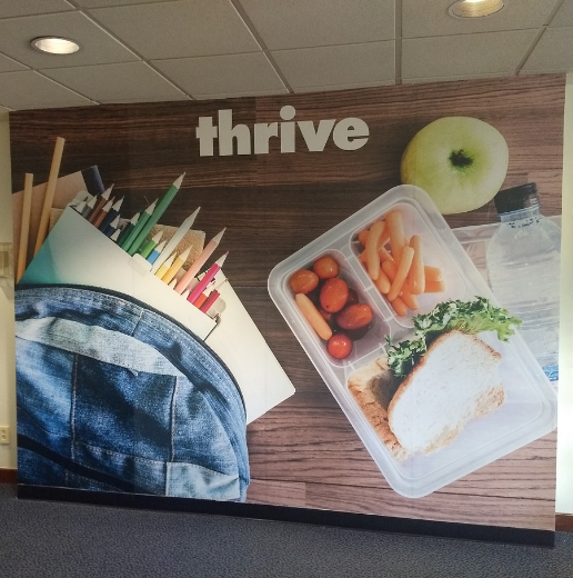 Promotional wall mural featuring a healthy lunch, colored pencils in a denim pouch, and a green apple with the word 'thrive'.