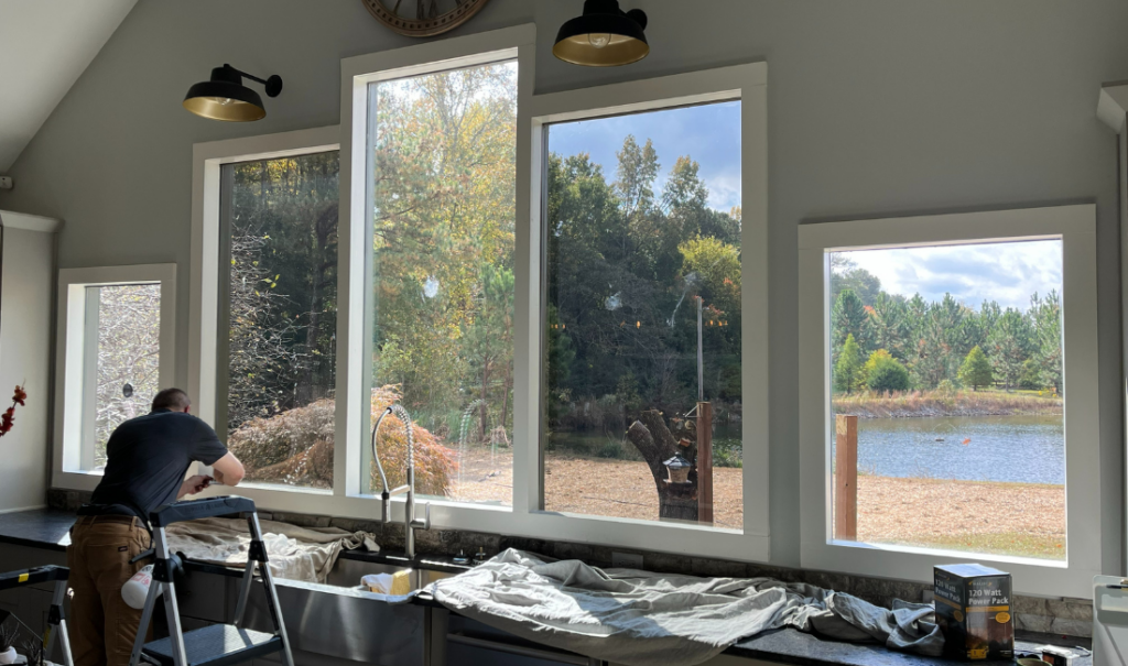 A professional installer from UHS Window Tinting is working on a home window tinting project near large windows overlooking a scenic view with trees and a pond.